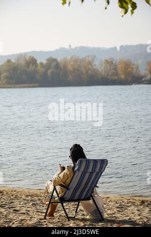 Das Mädchen mit schwarzen Haaren sitzt auf dem Ufer des Flusses auf dem Sand in einem Liegestuhl und schaut auf das Mobiltelefon Stockfoto