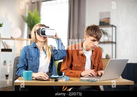 Glückliche junge Freunde, die am Tisch sitzen und moderne Geräte für Arbeit oder Studium verwenden. Hübsches blondes Mädchen mit VR-Brille und Spaß an der künstlichen Realität, während fokussierter Mann auf dem Laptop tippt Stockfoto