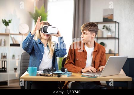 Glückliche junge Freunde, die am Tisch sitzen und moderne Geräte für Arbeit oder Studium verwenden. Hübsches blondes Mädchen mit VR-Brille und Spaß an der künstlichen Realität, während fokussierter Mann auf dem Laptop tippt Stockfoto