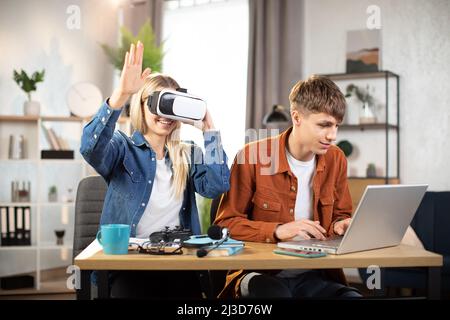 Glückliche junge Freunde, die am Tisch sitzen und moderne Geräte für Arbeit oder Studium verwenden. Hübsches blondes Mädchen mit VR-Brille und Spaß an der künstlichen Realität, während fokussierter Mann auf dem Laptop tippt Stockfoto