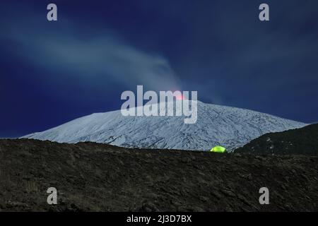 Beleuchtete Zelt am Rande eines alten Krater vor dem schneebedeckten Ätna, die vulkanischen Dampf emittieren, Ätna Park - Sizilien Stockfoto