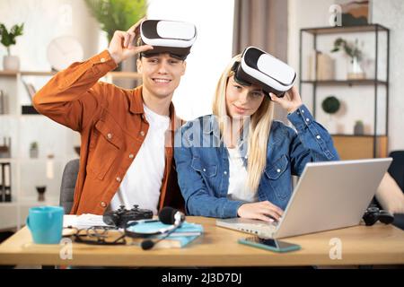 Junges Paar in Freizeitkleidung, das während der Freizeit zu Hause ein VR-Headset und einen Laptop verwendet, um Spiele zu spielen. Konzept von Familie und Unterhaltung. Stockfoto