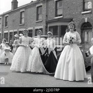 1961, historische Prozession am 1. Mai, halten junge Mädchen das schwere Gewebe des Zuges des Gewandes der frisch gekrönten May Queen der Stadt, während sie mit ihr in einer Straße mit viktorianischen Reihenhäusern, Stockport, Manchester, England, Großbritannien, für ein Foto stehen. Stockfoto