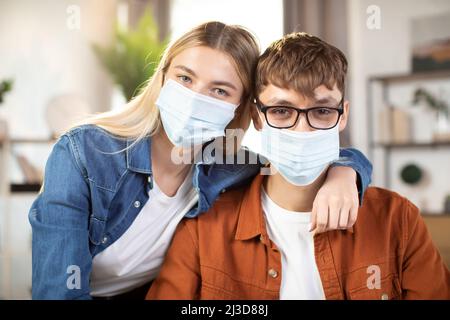 Studenten, Mann und Frau in zwanglosem Outfit und Gesichtsmasken posieren vor der Kamera, sitzen am Tisch und mit Laptop. Junge Partner, die von zu Hause aus zusammenarbeiten oder Remote online arbeiten Stockfoto