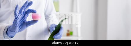 Beschnittene Ansicht des Laborassistenten in Latexhandschuhen mit Aloe-Blatt und Behälter mit Creme, Banner Stockfoto