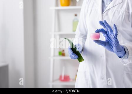 Beschnittene Ansicht des Laborhelfers in Latexhandschuhen mit Aloe-Blatt und Behälter mit Creme Stockfoto