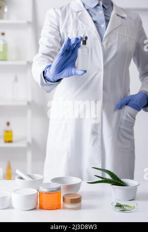 Abgeschnittene Ansicht des Laborassistenten in Latexhandschuhen, die eine Flasche mit Serum halten und mit der Hand in der Tasche stehen Stockfoto