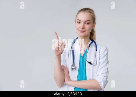 Junge europäische Frau mit Arzt und Stethoskop, die mit dem Finger nach oben zeigt, Geste, die aufpasst. Hochformat auf weißem Hintergrund Stockfoto