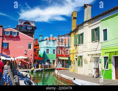 Venedig (Burano), Italien - Mai 9. 2019: Blick auf den idyllischen Wasserkanal mit Touristen, bunten Häusern, Restaurant, klarem blauen Himmel mit flauschigen Stockfoto