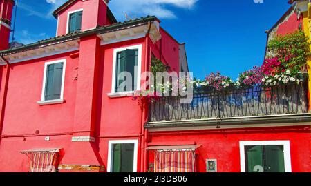 Venedig (Burano), Italien - Mai 9. 2019: Blick auf die hellrote Hausfassade im mediterranen Stil mit blumengeschmücktem Balkon gegen klares Blau Stockfoto