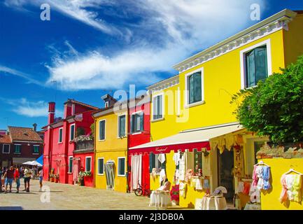Venedig (Burano), Italien - Mai 9. 2019: Blick auf den Platz mit leuchtend gelben, rot gefärbten Häusern, Modeboutique gegen klaren blauen Himmel, flauschige Wolken Stockfoto