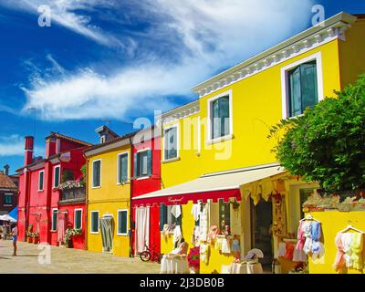 Venedig (Burano), Italien - Mai 9. 2019: Blick auf den Platz mit leuchtend gelben, rot gefärbten Häusern, Modeboutique gegen klaren blauen Himmel, flauschige Wolken Stockfoto