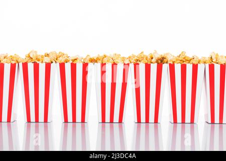 Kino-, Film- und Theaterkonzept. Popcorn-Boxen mit weißen und roten Streifen voller Süße mit Karamell-Popcorn auf weißem reflektierendem Hintergrund in Weiß Stockfoto