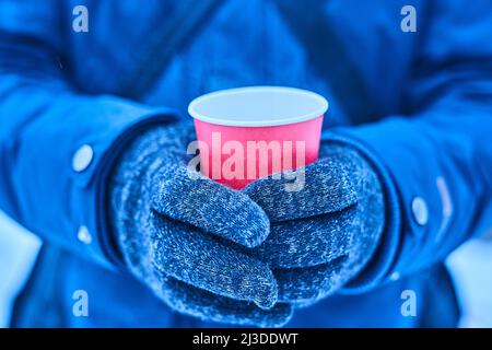 Becher in der Hand im Winter mit einem heißen Getränk, Modell für Ihren Text, Winterspaziergang mit heißem Tee in Handschuhen, roter Papierbecher eco auf blauem Hintergrund. Hochwertige Fotos Stockfoto