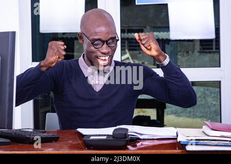 Ein gutaussehender Mann in einem blauen Poloshirt sitzt am Schreibtisch und schaut sich ein Dokument an und schreit vor Freude. Stockfoto