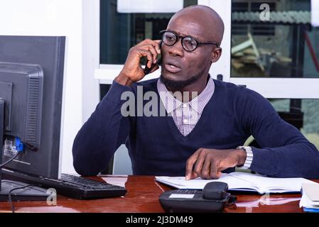 Ein hübscher Mann in einem blauen Poloshirt sitzt am Schreibtisch und redet am Handy. Stockfoto