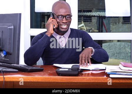 Ein hübscher Mann in einem blauen Poloshirt sitzt am Schreibtisch und redet lächelnd auf dem Handy. Stockfoto