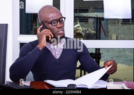 Ein hübscher Mann in einem blauen Poloshirt sitzt am Schreibtisch und redet am Handy, während er ein Dokument überprüft. Stockfoto