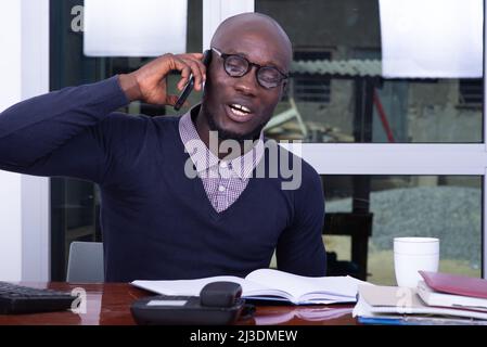Ein hübscher Mann in einem blauen Poloshirt sitzt am Schreibtisch und lacht am Handy. Stockfoto