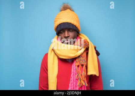 Afrikanischer lustiger Mann in mehreren Hüten und Schals, der traurig ist oder sich auf blauem Hintergrund kalt fühlt. Wintermode. Stockfoto