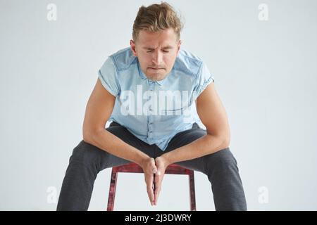 Brutaler Mann in einem Hemd mit kurzen Ärmeln auf einem roten Stuhl sitzend, seine Fäuste geballt, leicht gebeugt, unter dem Blick der Stirn, auf die Knie gelehnt Stockfoto
