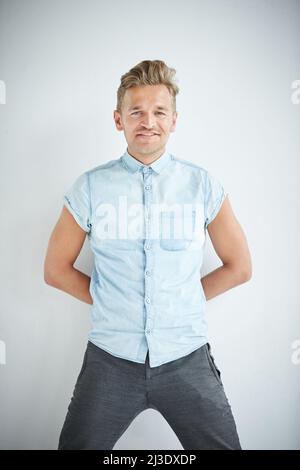 Brutaler Mann mit einem kleinen Bart lehnt sich an die Wand, trägt ein blaues Kurzarm-Hemd und eine graue Hose, die Hände sind hinter seinem Rücken Stockfoto