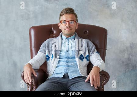 Porträt eines stilvollen intelligenten Mannes mit Brille starrt in die Kamera, gute Sicht, kleines unrasiert, charismatisches, blaues Hemd, Grauer Pullover, sitzend Stockfoto