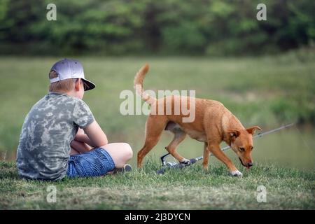 Naturforscher Junge mit Ingwer Hund sitzt in der Nähe des Teiches und wartet auf einen Fisch zu fangen. Geduldiger junger Fischer, der im Sommer die Natur mit Haustier genießt Stockfoto