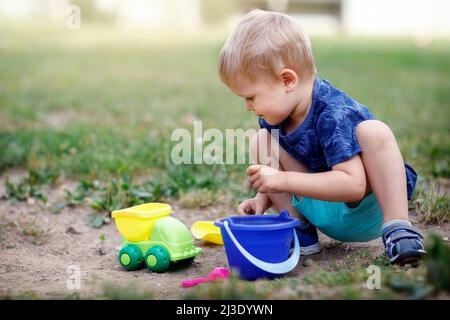 Kleiner Junge spielt auf der Wiese mit Sandspielzeug. Vorsichtig Sand in einen Eimer geben. Helle Farben und Sonnenschein. Freizeit in der Natur Stockfoto