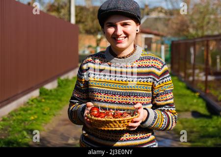 Unschärfe ukrainische junge lächelnde Frau in Hut hält Platte der Sammlung von rotem Ei auf Natur Hintergrund. Ostern, Ukraine. Bemalte Eier basteln. Pysanka Stockfoto