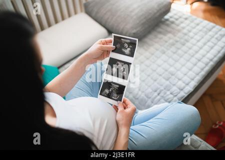 Eine schwarzhaarige kaukasische Schwangerin sieht sich Ultraschallbilder von Babys an. Sie hält Ultraschallpapier vor ihren schwanger werdenden Bauch. Eine Frau sitzt. Stockfoto