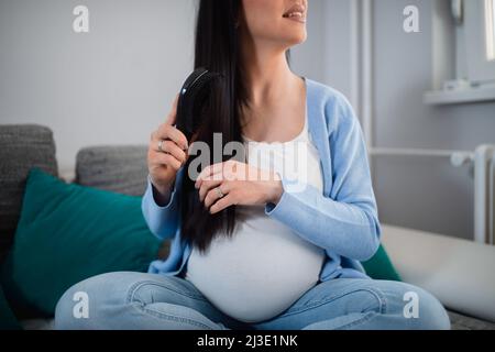 Lange schwarzhaarige, schwanger kaukasische Kämme ihre Haare mit einer schwarzen Haarbürste, während sie mit gekreuzten Beinen auf dem Sofa sitzt und zur Seite schaut. Stockfoto