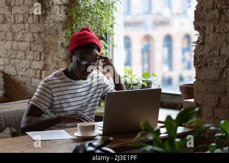 Nachdenklicher junger afroamerikanischer Freiberufler mit Brille, der im Café auf dem Laptop arbeitet Stockfoto
