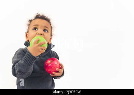 Liebenswerter afroamerikanischer kleiner Junge mit lockigem Haar, der einen grünen Apfel isst und einen roten Apfel im Studio hält, weißer Hintergrund, mittlerer Nahaufnahme, Kopierraum. Hochwertige Fotos Stockfoto