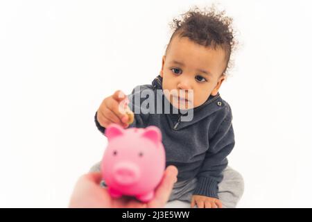 Niedlicher afroamerikanischer kleiner Junge, der auf dem Boden sitzt und eine Münze in das rosa Sparschwein mit einem ernsthaften Gesicht einlegt, Studio Shot Medium Full Shot White Background copy space. Hochwertige Fotos Stockfoto