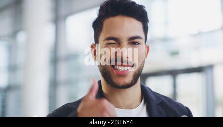Es ist ein großartiger Tag für Geschäftsreisende. Eine kurze Aufnahme eines jungen Geschäftsmanns, der beim Gehen durch ein modernes Büro zwinkert und Daumen nach oben zeigt. Stockfoto