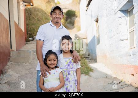 Latino Vater mit seinen zwei glücklichen Töchtern vor seinem Haus in ländlicher Umgebung-hispanischen Vater umarmt seine Töchter Stockfoto