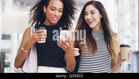 Sie teilen alles. Eine kurze Aufnahme von zwei jungen Geschäftsfrauen, die mit einem Smartphone chatten und während einer Kaffeepause in einem Büro spazieren gehen. Stockfoto