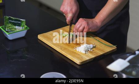 Der Koch schneidet grüne Zwiebeln. KUNST. Männliche Hände im Rahmen schneiden grüne Zwiebeln mit einem Messer auf dem Brett. Auf dem Tisch sind Brett, Zwiebel Stockfoto