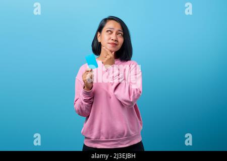 Porträt einer jungen asiatischen Frau mit Kreditkarte und seitlichem Blick mit zweifelhaftem und skeptischem Ausdruck Stockfoto