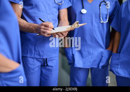 Überprüfung der Patientenakte vor der Operation. Ausgeschnittene Aufnahme eines Arztes, der mit Kollegen Notizen auf ein Klemmbrett schreibt Stockfoto