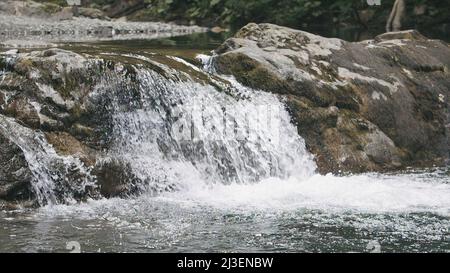 Schöner natürlicher Wasserfall des Flusses im Wald. Kreativ. Der Flussbach verwandelt sich in einen Wasserfall und fließt weiter. Steinlandschaft von Flussformen Stockfoto
