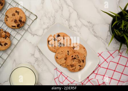 Schokoladenkekse auf einem weißen Teller mit einem Glas Milch, frisch gebackenen Keksen und Dekor auf Marmorhintergrund. Bäckerei, Konditorei Konzept Stockfoto