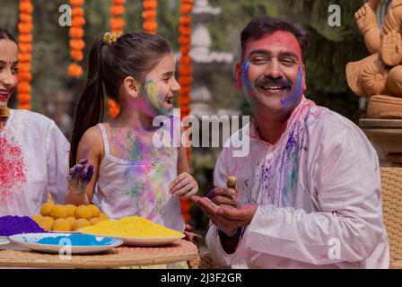 Enkelin, die Farbe auf das Gesicht des Großvaters anwendet, und Mutter, die anläßlich von Holi daneben sitzt Stockfoto