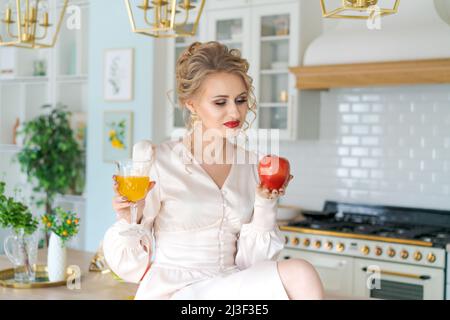 Schöne erfolgreiche Frau genießen frischen Orangensaft. In ihrer Küche trinkt die Dame frisch zubereitete Smoothie voller Vitamine. Hält Glas und roten Apfel in der Hand. Gesunder Lebensstil Stockfoto