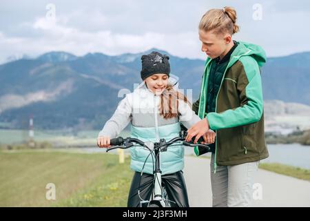 Bruder erklärt seiner Schwester die Grundlagen eines Fahrradfahrens, das ihr hilft und lehrt, erste Schritte im Fahrradfahren zu machen. Happy Outdoor Kindheit Konzept Bild. Stockfoto