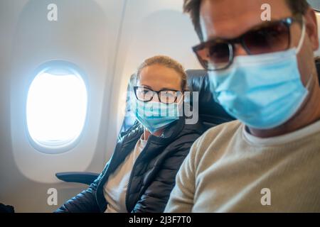 Ein junges Paar, das Gesichtsmasken trägt, während es im Flugzeug unterwegs ist. Neuer Normalweg nach dem covid-19-Pandemiekonzept Stockfoto