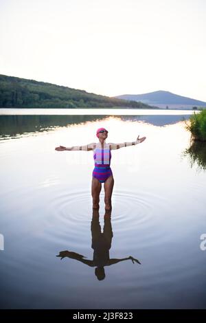 Porträt einer aktiven älteren Schwimmerin, die im Freien im See steht und sich ausdehnt. Stockfoto