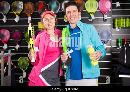 Lächelnde Frau mit Mann stehen in Sportbekleidung und halten moderne Padel-Schläger und -Kugeln im Laden Stockfoto