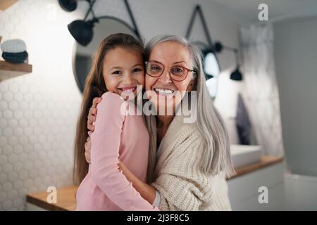 Ältere Großmutter und Enkelin stehen drinnen im Badezimmer, umarmen und betrachten die Kamera. Stockfoto